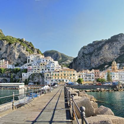 Panoramisch uitzicht op Amalfi vanaf een stenen pier, met kleurrijke gebouwen tussen dramatische kliffen en de Middellandse Zee.