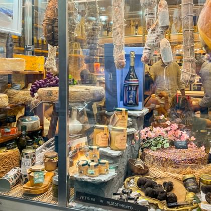 Etalage in Ligurië met Italiaanse specialiteiten: hangende salami, kaaswielen, wijn, truffels en bloemen op gelaagde planken.