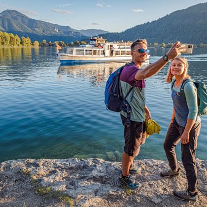 Twee wandelaars met rugzakken maken een selfie op rotsachtige oever van Tegernsee meer, met passagiersboot en bergen op achtergrond.
