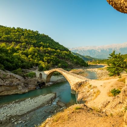 Historische Kadius stenen brug over de turquoise Vjosa-rivier in Hidden Valley van Albanië, met groene bergen en rotsachtige kliffen onder blauwe lucht.