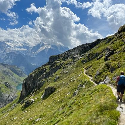 Reto wandelt met blauwe rugzak op bergpad van de Haute Route naar Lac de Louvie, omringd door groene alpenhellingen en besneeuwde toppen.