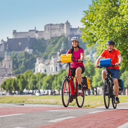 Twee fietsers met fietstassen op rood fietspad langs de Salzach in Salzburg met vesting Hohensalzburg op de heuvel.