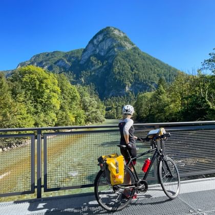 Fietser met helm op Saalach brug met toerfiets, uitzicht op rivier met Muellnerhorn berg op achtergrond onder blauwe lucht.