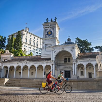 Twee fietsers rijden langs de historische klokkentoren en zuilengalerij op Piazza della Libertà in Udine, Friuli.