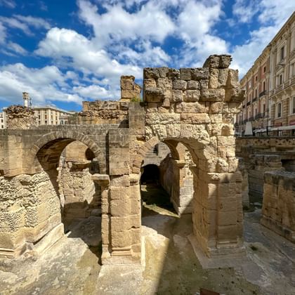 Oude stenen ruïnes van een gotische boogstructuur in Lecce's Romeins amfitheater, omringd door historische gebouwen onder een blauwe hemel.