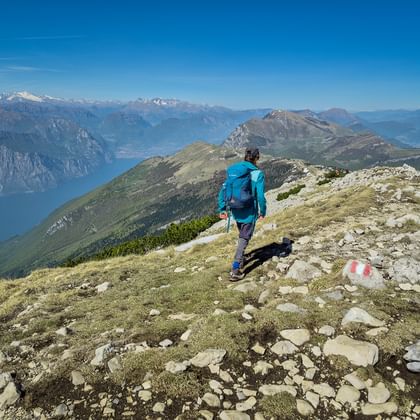 Wandelaar in blauwe jas op Monte Baldo bergpad met uitzicht op Gardameer en besneeuwde Alpen onder heldere blauwe lucht.