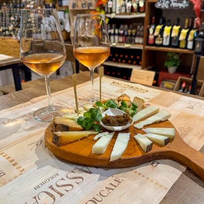 Houten kaasplank met diverse kazen, groenten en chutney, vergezeld van drie glazen witte wijn op een tafel in een wijnwinkel.