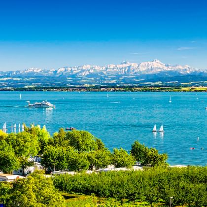 Uitzicht op het Bodenmeer vanuit Hagnau met zeilboten op blauw water, groene wijngaarden op de voorgrond en besneeuwde Alpen op de achtergrond.