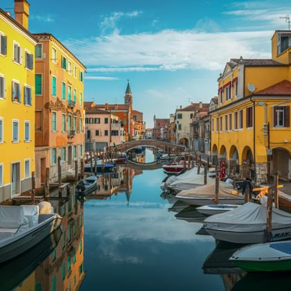Canale Vena in Chioggia met kleurrijke huizen in oranje en geel, afgemeerde boten en een brug. Kerktoren zichtbaar op de achtergrond.