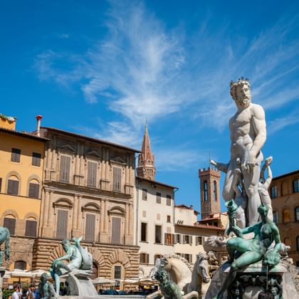 Neptunusfontein in Florence met wit marmeren beeld en bronzen figuren, omringd door historische gebouwen en kerktorens onder blauwe lucht.