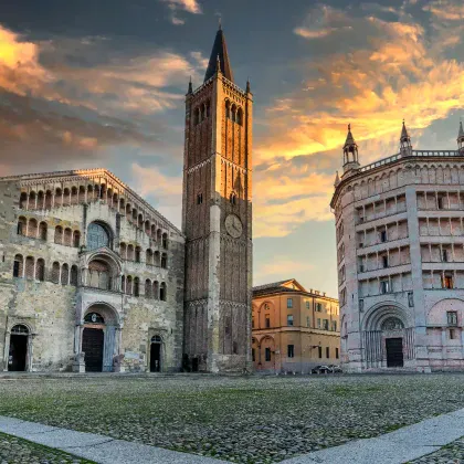 Duomo di Parma kathedraalcomplex met Romaanse gevel, hoge klokkentoren en achthoekig baptisterium onder dramatische zonsonderganghemel in Emilia.