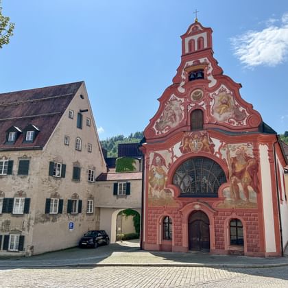 Barokke Heilig-Geist-Spitalkirche in Füssen met sierlijke rode gevel met religieuze fresco's en klokkentoren, omgeven door traditionele gebouwen.