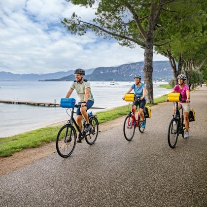 Drie fietsers met fietstassen rijden langs een met bomen omzoomd pad aan het Gardameer. Bergen zichtbaar over het kalme blauwe water.