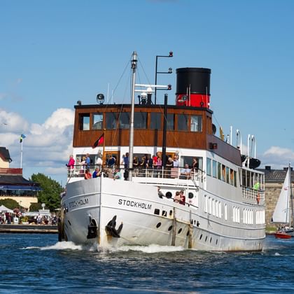 Witte stoomboot Stockholm met passagiers aan dek varend in de haven van Vaxholm. Historische gebouwen en vestingmuren zichtbaar aan de kust.