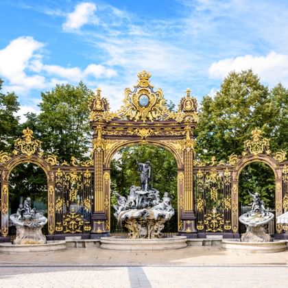Sierlijke vergulde smeedijzeren hekken met fonteinen op Place Stanislas in Nancy, met barokke decoratieve elementen onder blauwe lucht met bomen.