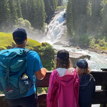 Familie van drie personen staat op houten uitkijkplatform en kijkt naar Krimml waterval in weelderig groen bos.