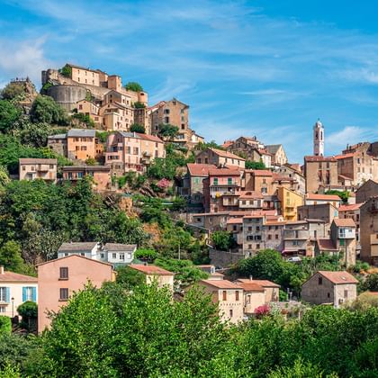 Bergstad Corte op Corsica met kleurrijke stenen gebouwen die zich uitstrekken over bergachtig terrein, omgeven door weelderige groene vegetatie.
