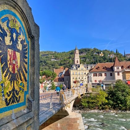 Postbrug in Merano met kleurrijk heraldisch adelaarsmozaïek. De brug overspant een rivier met historische gebouwen en heuvels op de achtergrond.
