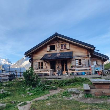 Traditionele houten berghut Cab de Louvie met zonnepanelen op het dak, gelegen op grasrijke helling met besneeuwde Alpentoppen op de achtergrond.