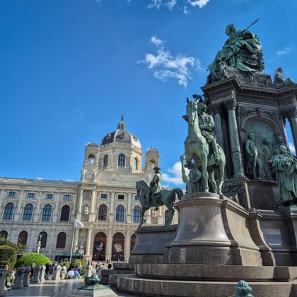 Maria Theresia-standbeeld op hoog monument met bronzen figuren en paarden, met het Kunsthistorisches Museum op de achtergrond onder blauwe lucht.