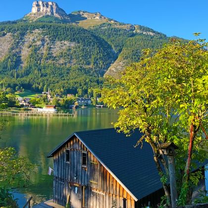 Uitzicht op het Altaussee meer met houten boothuis, groene bomen en dramatische bergtoppen onder heldere blauwe hemel in de Alpen.