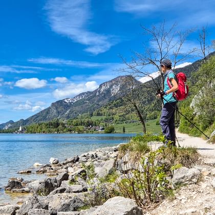 Vrouwelijke wandelaar met rode rugzak op rotsachtige oever van Wolfgangsee in Salzkammergut, Oostenrijk, met bergen en blauwe lucht.
