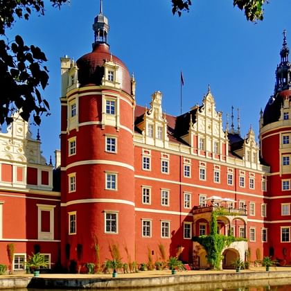 Rood barok kasteel met witte decoratieve elementen en ronde torens in Bad Muskau, omgeven door water en groene bomen onder blauwe lucht.