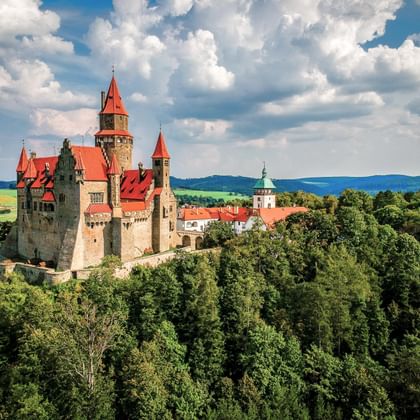 Kasteel Bouzov met rode daken en torens op een beboste heuvel in Tsjechië, omringd door groene bomen onder een bewolkte blauwe lucht.