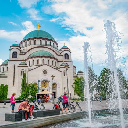 Belgrado Kathedraal met witte muren en groene koepels met gouden kruisen. Fonteinstralen spuiten op voorgrond met bezoekers dichtbij.