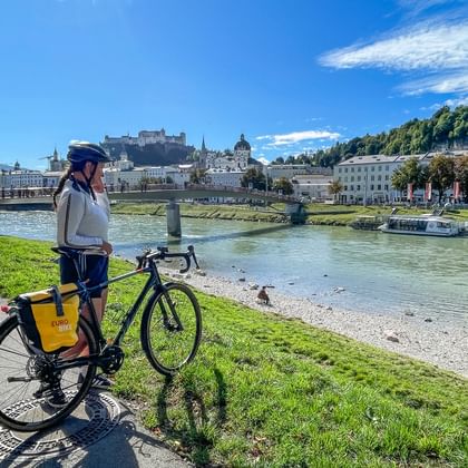 Fietser met helm staat met fiets op groene oever van de Salzach in Salzburg, met historisch stadscentrum en fort zichtbaar.