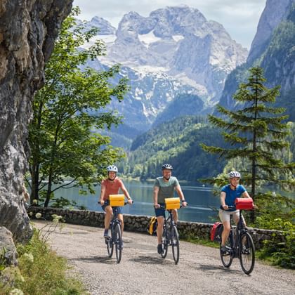 Drie fietsers rijden langs het Gosausee-meer met dramatische Dachstein-bergtoppen op de achtergrond. Rotswand links, alpenmeer en bos zichtbaar.