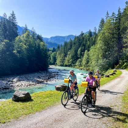 Drie fietsers op een pad langs de turquoise Saalach bij Lofer, omringd door dichte bossen en bergen onder een blauwe hemel.