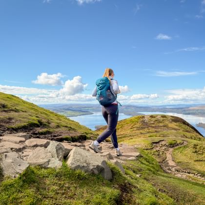 Vrouwelijke wandelaar met rugzak op rotsachtig pad op groene heuvel met uitzicht op Loch Lomond onder blauwe lucht met witte wolken.