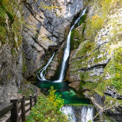 Savica-waterval die over met mos bedekte rotsen naar beneden stroomt in een smaragdgroene poel in Slovenië. Een houten uitkijkplatform is zichtbaar op de voorgrond.