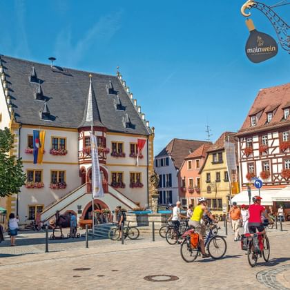 Fietsers op het marktplein van Volkach met wit stadhuis met rode trap, vakwerkhuizen en sierlijk Mainwein-bord onder blauwe lucht.