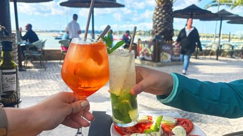Twee handen proosten met cocktails in een strandrestaurant in de Algarve. Oranje en groene drankjes met rietjes, tomaat-mozzarella bord zichtbaar.