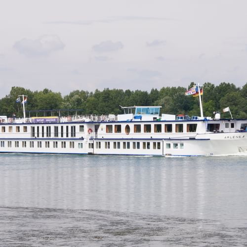 Wit riviercruiseschip MS De Willemstad varend op kalm water met beboste kustlijn op de achtergrond onder bewolkte hemel.