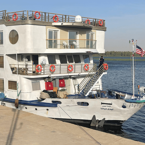 Wit Nebu riviercruiseschip met blauwe rand aangemeerd aan de Nijl. Het schip heeft meerdere dekken met balkons en een Amerikaanse vlag achteraan.