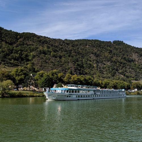 MS Switserland riviercruiseschip met witte romp en blauw bovendek varend op kalm groen water met beboste heuvels en dorp op de achtergrond.