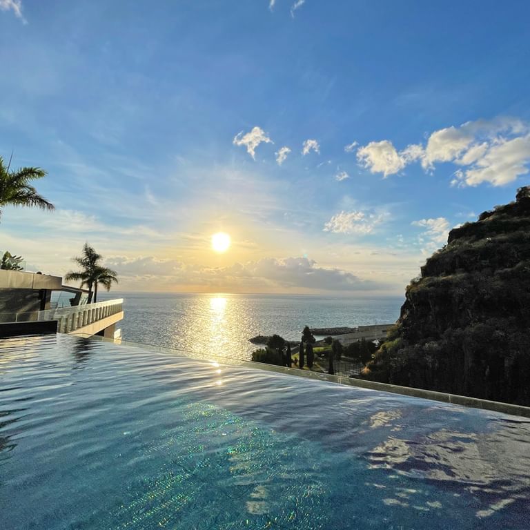 Infinity pool van Hotel Savoy Saccharum in Calheta met oceaanzicht bij zonsondergang. Palmbomen en rotsklif zichtbaar, gouden zon weerkaatst in water.