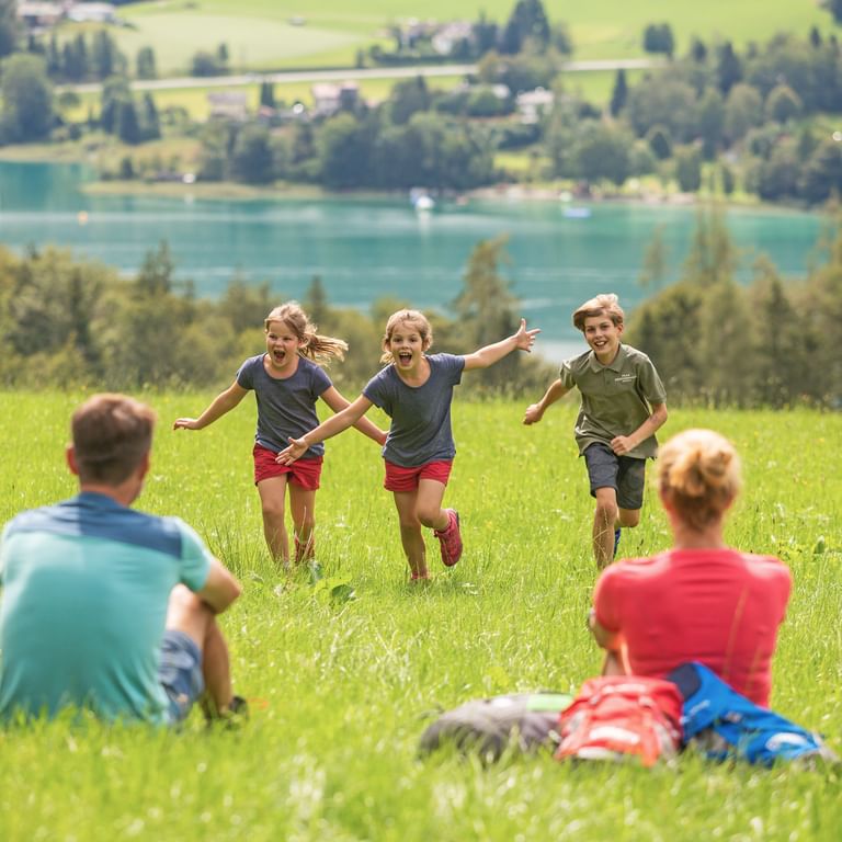Drie kinderen rennen door groene weide naar ouders tijdens picknick. Turquoise alpenmeer en glooiende heuvels op achtergrond.