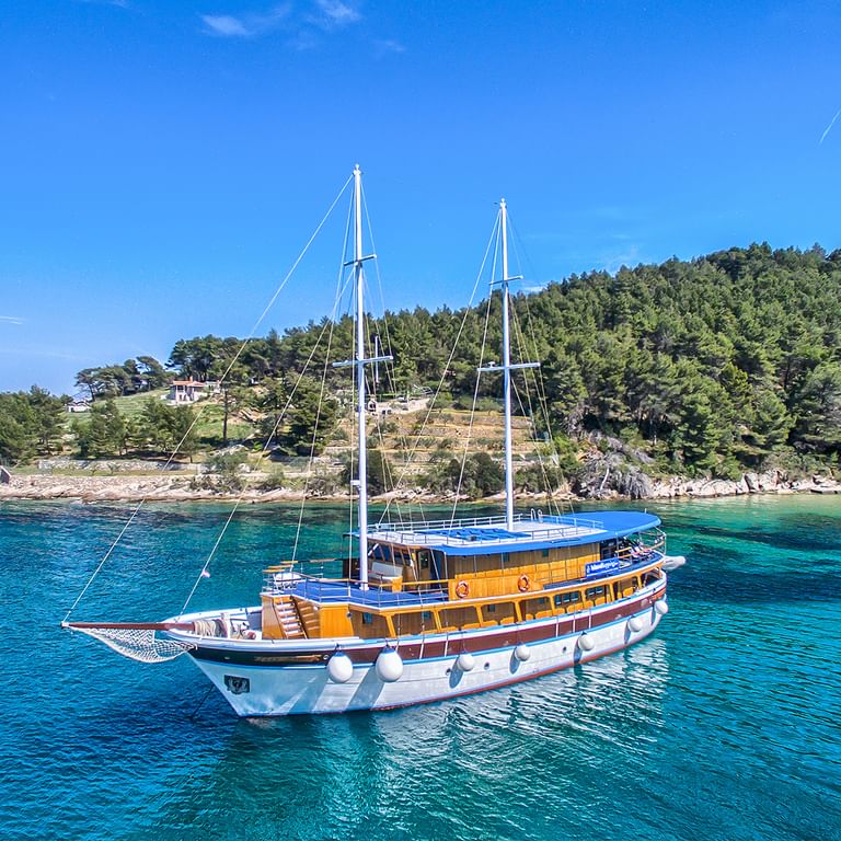 Tweemast zeilschip San Snova met witte en houten romp voor anker in turquoise Adriatische wateren nabij beboste Kroatische kustlijn.